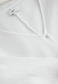 Haut blanc texturé avec un décolleté en V, doté d'un petit bouton et de détails cousus, fabriqué en tissu léger. L'étiquette indique "NEXT."