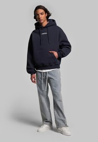 Hoodie bleu marine avec cordons de serrage et poche avant, associé à un jean en denim clair. Comprend un texte de logo blanc et des baskets blanches décontractées.