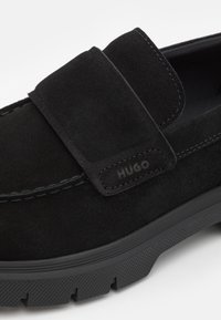 Mocassins en daim noir avec une large sangle, des détails cousus et une semelle en caoutchouc texturée avec un logo de la marque sur le côté.