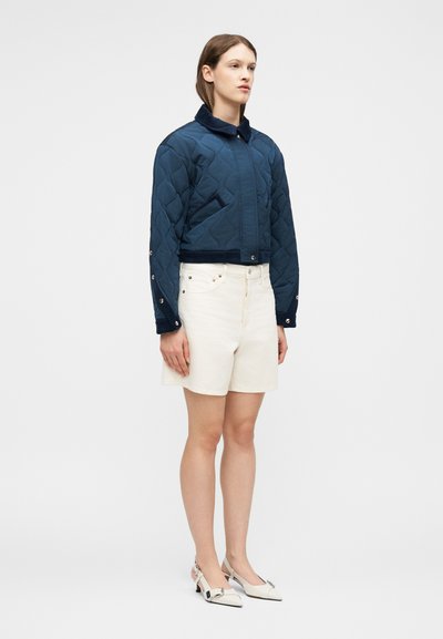Femme portant une veste matelassée bleu marine avec des boutons-pression et un short blanc taille haute, debout devant un fond clair uni.