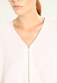 V-ringad tröja i ljusrosa, tillverkad av strukturerad tyg. Har en framdragkedja och en minimalistisk design. Slät yta på kragen.