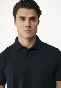 Svart polo skjorta i mjukt tyg, med krage, knappslå med tre knappar och diskret logotyp i matchande nyans på bröstet.