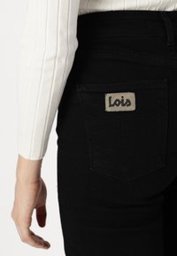Jeans in denim nero con una tasca posteriore prominente che presenta un'etichetta beige impresso con "Lois". Il tessuto ha una texture liscia e opaca.