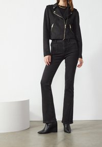 Giacca cropped in suede sintetico nera con cerniere dorate, abbinata a jeans neri a vita alta con gamba a zampa e stivaletti neri con zeppa alla caviglia. Texture liscia.