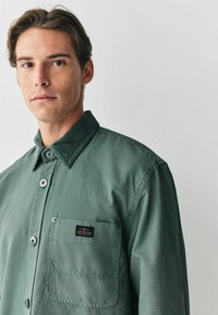 Grünes Hemd mit Knöpfen und einem Cordkragen, ausgestattet mit einer Brusttasche und einem Markenetikett. Der Stoff hat eine glatte Textur und eine lässige Passform.