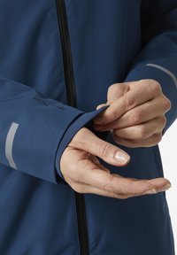 Navyblauwe softshell jas met rits, met reflecterende strepen op de manchet en een close-up van een hand die de mouw aanpast.