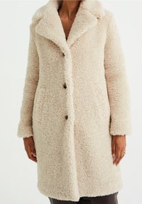 Abrigo de piel sintética beige con cuello notch, cierre de botones y bolsillos laterales. Tejido suave y texturizado con un corte relajado y mangas largas.