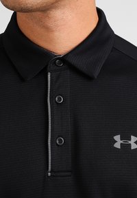 Camisa polo negra con tejido texturizado, cuello abotonado, acentos grises a lo largo de la tapeta y un pequeño logo en el pecho.