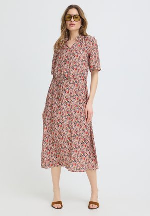 Moteris, dėvinti geltonai tonuotus saulės akinius ir gėlėtą midi suknelę su trumpomis rankovėmis, stovinti prieš paprastą šviesią foną.