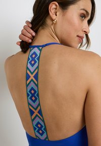 Costume da bagno blu con schiena bassa e un motivo geometrico colorato in viola, verde e arancione sulle spalline. Tessuto morbido.