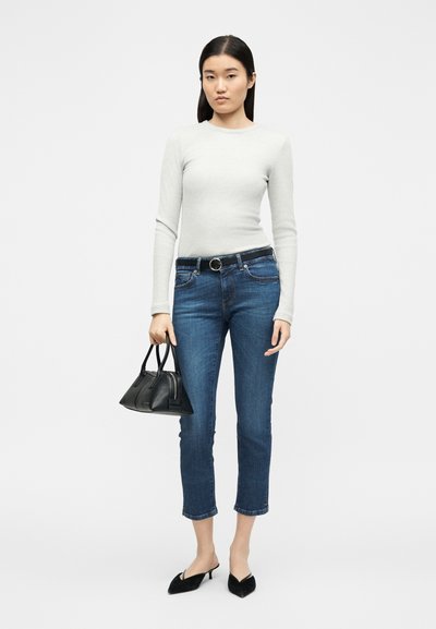 Femme portant un haut à manches longues gris clair, jean bleu court avec ceinture noire, escarpins noirs à talons bas, tenant un sac à main noir sur fond blanc.