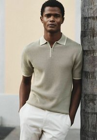 Un jeune homme portant un polo en maille beige à manches courtes et un pantalon blanc, debout les mains dans les poches à côté d'un tronc d'arbre texturé.