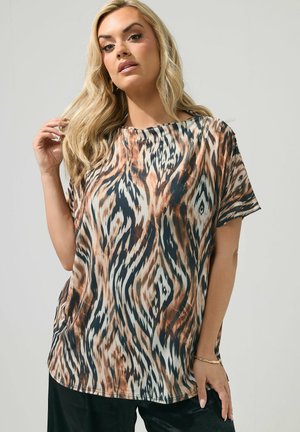 Kvinde med langt blond hår iført en løs top med korte ærmer med brun, sort og hvid abstrakt dyreprint, poserer mod en neutral baggrund.