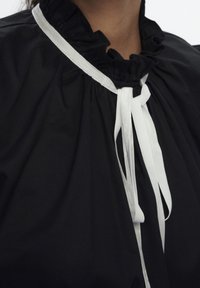 Zwarte stoffen blouse met een rimpelachtige halslijn en een witte satijnen strik, wat zorgt voor een samengevoegd en textuurachtig uiterlijk.