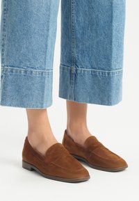 Bruine suède loafers met een platte zool, voorzien van gestikte details en een ronde neus, gecombineerd met lichtblauwe, wijdvallende, cropped denim broek.