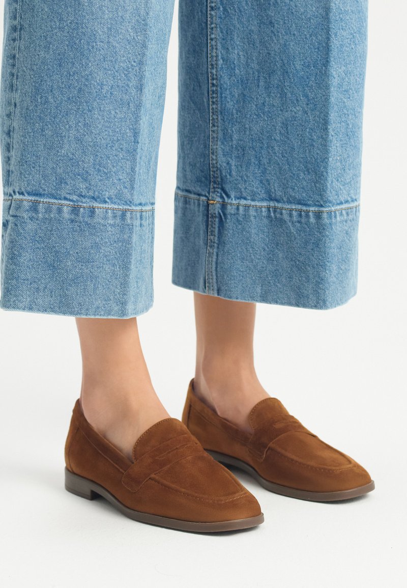 Bruine suède loafers met een platte zool, voorzien van gestikte details en een ronde neus, gecombineerd met lichtblauwe, wijdvallende, cropped denim broek.