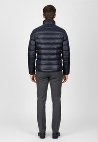 Marineblå pufferjakke med quiltet design, høj krave og lange ærmer, parret med grå bukser og mørke sko, set fra ryggen.