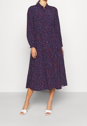 Sukienka na długi rękaw, zapinana na guziki, w kolorze granatowym z kolorowym wzorem liści. Zwiewna tkanina z talią podkreśloną paskiem, długość do połowy łydki, w zestawieniu z białymi sandałami.