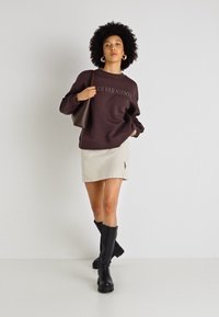 Sweatshirt castanho com o texto "DOLCE FAR NIENTE", saia mini canelada em creme, botas negras até ao joelho e uma mala de ombro em tom areia. Design e texturas simples.