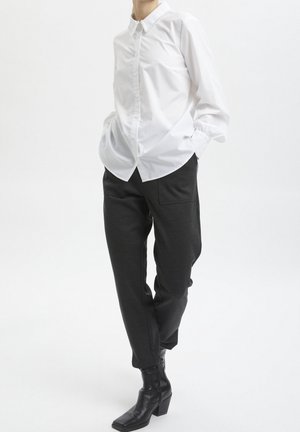 Personne portant une chemise blanche à manches longues boutonnée avec un col décoratif, un pantalon noir fuselé et des bottines noires à talons.