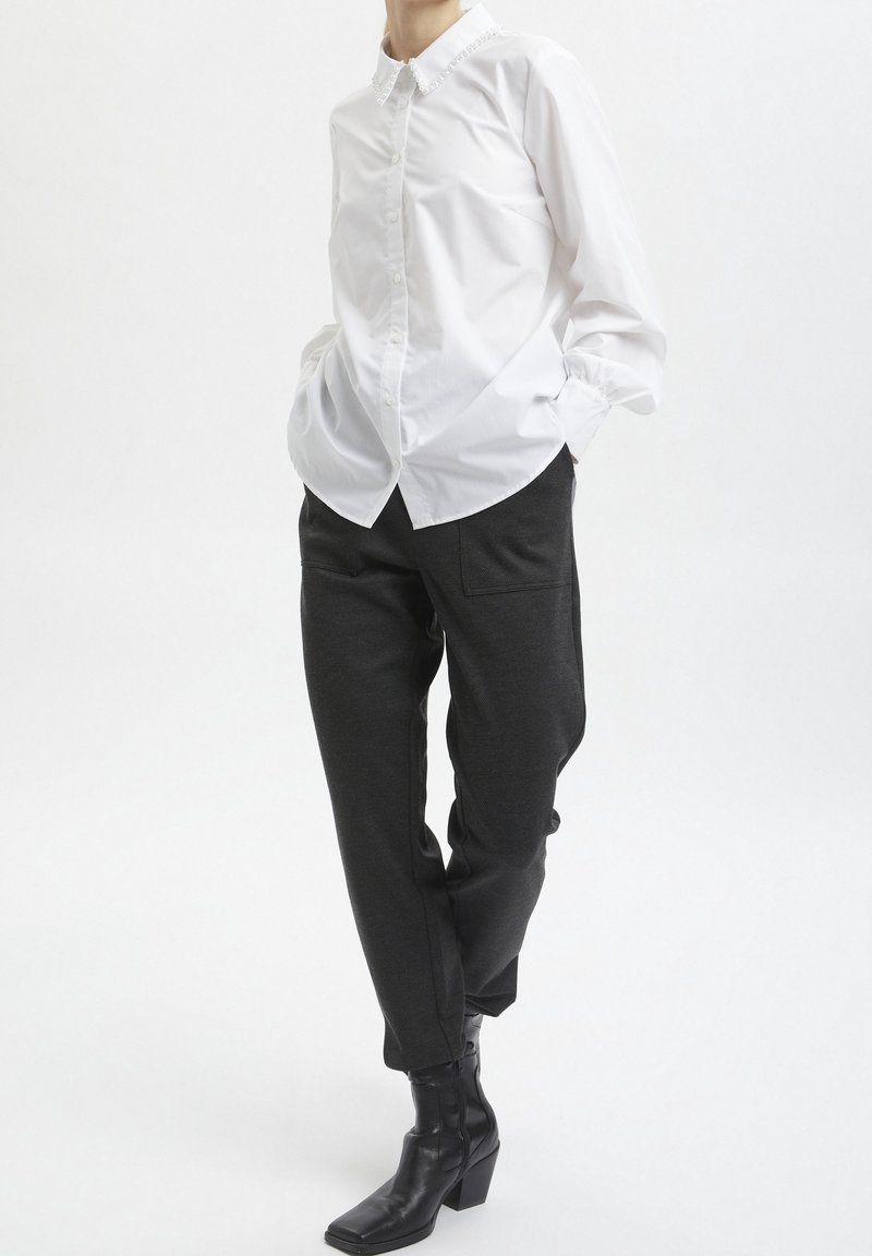 Personne portant une chemise blanche à manches longues boutonnée avec un col décoratif, un pantalon noir fuselé et des bottines noires à talons.