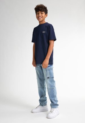 Granatowa koszulka z krótkim rękawem z kieszonką, w zestawie z jasnoniebieskimi spodniami cargo z kieszeniami i białymi sneakersami.