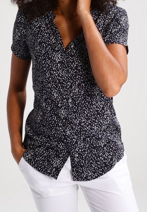 Femme portant une chemise noire à manches courtes à boutons avec des taches blanches et un pantalon blanc, une main dans la poche et l'autre près du visage.