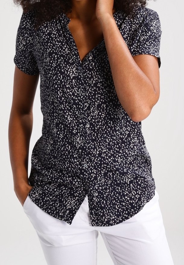 Femme portant une chemise noire à manches courtes à boutons avec des taches blanches et un pantalon blanc, une main dans la poche et l'autre près du visage.