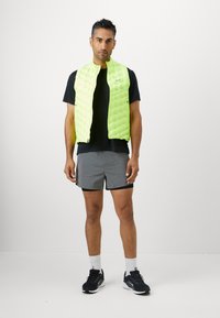 Gilet trapuntato verde neon con zip frontale, indossato sopra una maglietta nera. Abbinato a pantaloni corti grigi e scarpe da ginnastica nere.