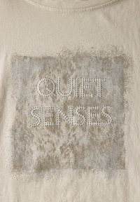 Béžové bavlněné tričko s texturovanou stříbrnou grafikou a nápisem "QUIET SENSES" zdobeným krystaly, orámované vybledlým vzorem.