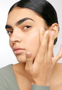 Nätt close-up av en person som applicerar en hudvårdsprodukt på sin kind. Huden är jämn, medeltonad, med glansiga läppar. Håret är mörkt och bakåtslickat.