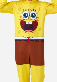 Pijama de felpa amarillo con la cara de un personaje de dibujos animados, con la parte inferior marrón, cremallera frontal y grandes ojos azules con detalles en negro.