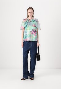 Bloemen top met een doorschijnende, transparante cape, in tinten groen, roze en blauw; gecombineerd met losse blauwe jeans en een zwarte handtas.