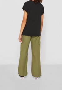 Osoba ubrana w luźne oliwkowe spodnie cargo, czarny krótki top i czarno-białe sneakersy, zwrócona plecami w stronę jednolitego tła.