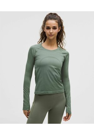 Femme aux cheveux bouclés attachés, portant un haut de sport sage vert ajusté à manches longues et un legging assorti, sur un fond uni.