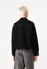 Jaqueta de denim preta, cropped, com detalhes em costura branca, punhos com botão e colarinho clássico. Combinada com jeans cinza de cintura alta.