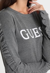 Pull gris avec encolure côtelée, arborant le texte blanc "GUES" et des accents à volants sur les manches, mettant en valeur la texture et les détails du design.
