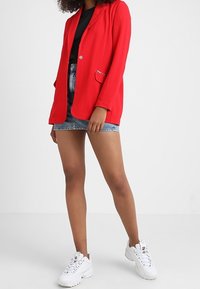 Femme portant un blazer rouge, un haut noir, une mini jupe en jean bleu et des baskets blanches sur un fond blanc uni.