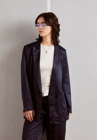 Blazer satinat bleumarin cu rever înclinat, închidere cu două butoane și buzunare laterale, purtat peste un top alb ajustat cu textură în dungi și pantaloni asortați.