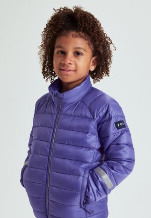 Nuori kiharatukkainen henkilö, jolla on violetti toppatakki heijastavilla nauhoilla, kädet taskuissa, yksinkertaista vaaleaa taustaa vasten.