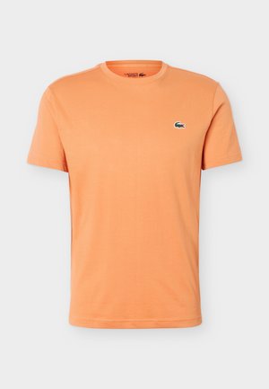 Πορτοκαλί κοντομάνικη μπλούζα με στρογγυλή λαιμόκοψη και μικρό πράσινο λογότυπο κροκοδείλου της Lacoste στο αριστερό στήθος, μονόχρωμο σχέδιο σε λευκό φόντο.