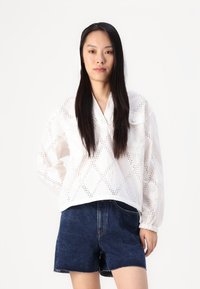 Vit broderad blus med diamantformade utskärningar, långa ärmar och avslappnad passform, parat med mörkblå denimshorts.