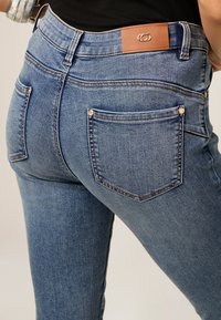 Jeans ajustados de mezclilla, en un lavado azul medio. Incluyen un parche de cuero marrón con el logo y dos bolsillos traseros con herrajes plateados.