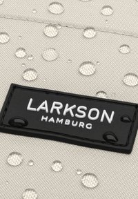 Gouttes d'eau sur un tissu clair entourant une étiquette de marque rectangulaire noire Larkson Hamburg cousue sur le matériau.