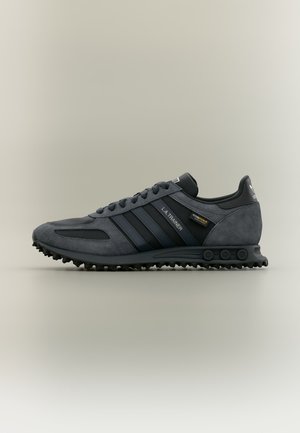 Σκούρο γκρι αθλητικό παπούτσι Adidas L.A. Trainer με μαύρες ρίγες, πάνελ από σουέτ και δίχτυ, κορδόνια και σκληρή σόλα με καρφιά, εμφανίζεται στο πλάι σε ουδέτερο φόντο.