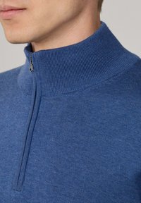 Pull en tricot bleu à col zippé haut, texture lisse et détails de couture minimaux près de la fermeture éclair.