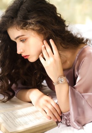 Montre dorée avec un bracelet en maille et un cadran circulaire texturé repose sur un poignet. La personne a des cheveux ondulés et foncés, portant un haut violet clair.