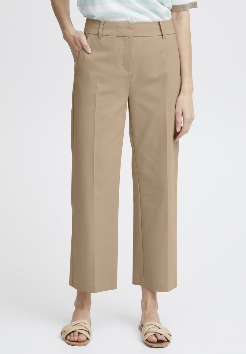 Pantalons beige à jambes larges avec une texture lisse, un léger éclat et des poches avant. Design court mettant en valeur des sandales confortables.