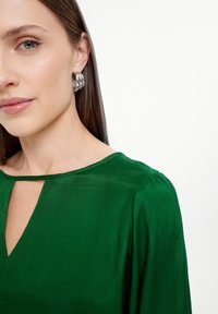 Zelena bluzica s laganim sjajem, okruglim izrezom i detaljem u obliku ključa; uparena sa srebrnim okruglim naušnicama. Glatka tekstura, duži rukavi.
