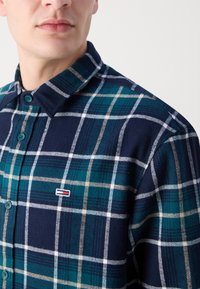 Camisa de franela con un patrón de cuadros en azul marino y azul verdoso, con acentos en blanco y beige. El cuello y los detalles de los botones son visibles, junto con un parche de logo.